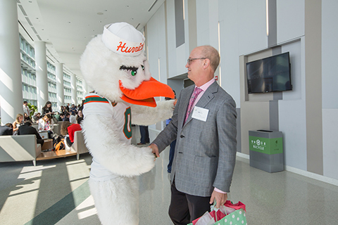 Man in a grey suit and pink shakes the hand of University of Miami mascot, Sebastian the Ibis